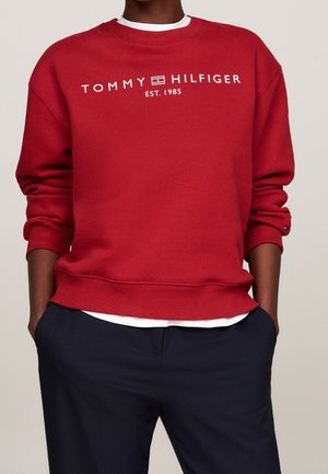 Osoba ubrana w czerwoną bluzę Tommy Hilfiger z białym logo oraz granatowe spodnie, ręce w kieszeniach, jednolite jasne tło.