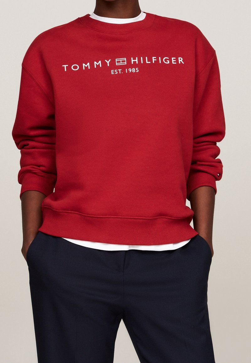 Personne portant un sweat-shirt rouge Tommy Hilfiger avec un logo blanc et un pantalon bleu marine, mains dans les poches, fond clair uni.
