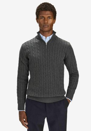 Mand iført en mørkegrå kabelstrikket kvart-zip sweater over en lyseblå skjorte med knapper og mørke bukser, stående med den ene hånd i lommen.