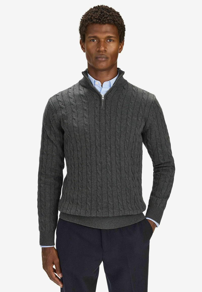 Homme portant un pull gris foncé en maille torsadée à fermeture éclair partielle sur une chemise bleu clair à boutons et un pantalon foncé, debout, une main dans la poche.