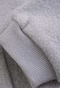 Grijze trui stof met een zachte, fuzzy textuur. Ribbenmanchet detail aan de onderkant heeft een gladde afwerking die het algehele ontwerp aanvult.