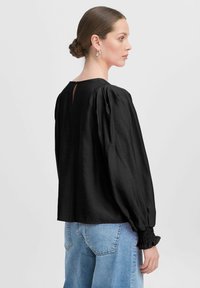 Blusa nera con maniche lunghe, spalle a sbuffo, schiena con apertura a forma di chiave e polsini arricciati. Abbinata a jeans azzurri a gamba larga.