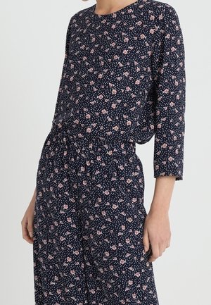 Frau trägt einen marineblauen Jumpsuit mit pinkem Blumenmuster und Dreiviertelärmeln. Sie steht mit einer Hand locker an der Seite.