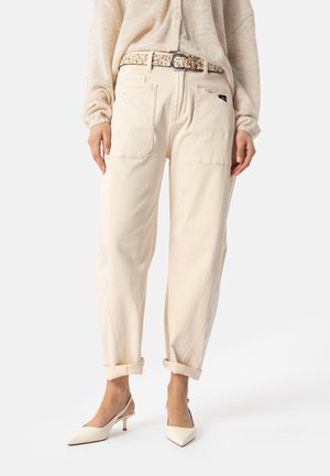 Frau trägt beige hoch taillierte Hose mit Vordertaschen, hochgekrempelten Bündchen, gemustertem Gürtel, cremefarbenem Pullover und beigen spitzen Slingback-Absätzen.