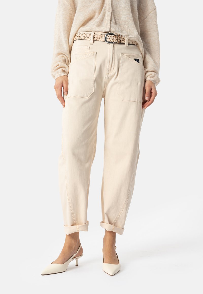 Femme portant un pantalon beige taille haute avec poches avant, revers roulés, ceinture à motif, pull crème et escarpins slingback beiges pointus.