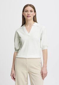 Pullover lavorato a maglia verde chiaro con scollo a V e maniche corte a sbuffo, abbinato a pantaloni beige. Texture morbida e vestibilità rilassata.