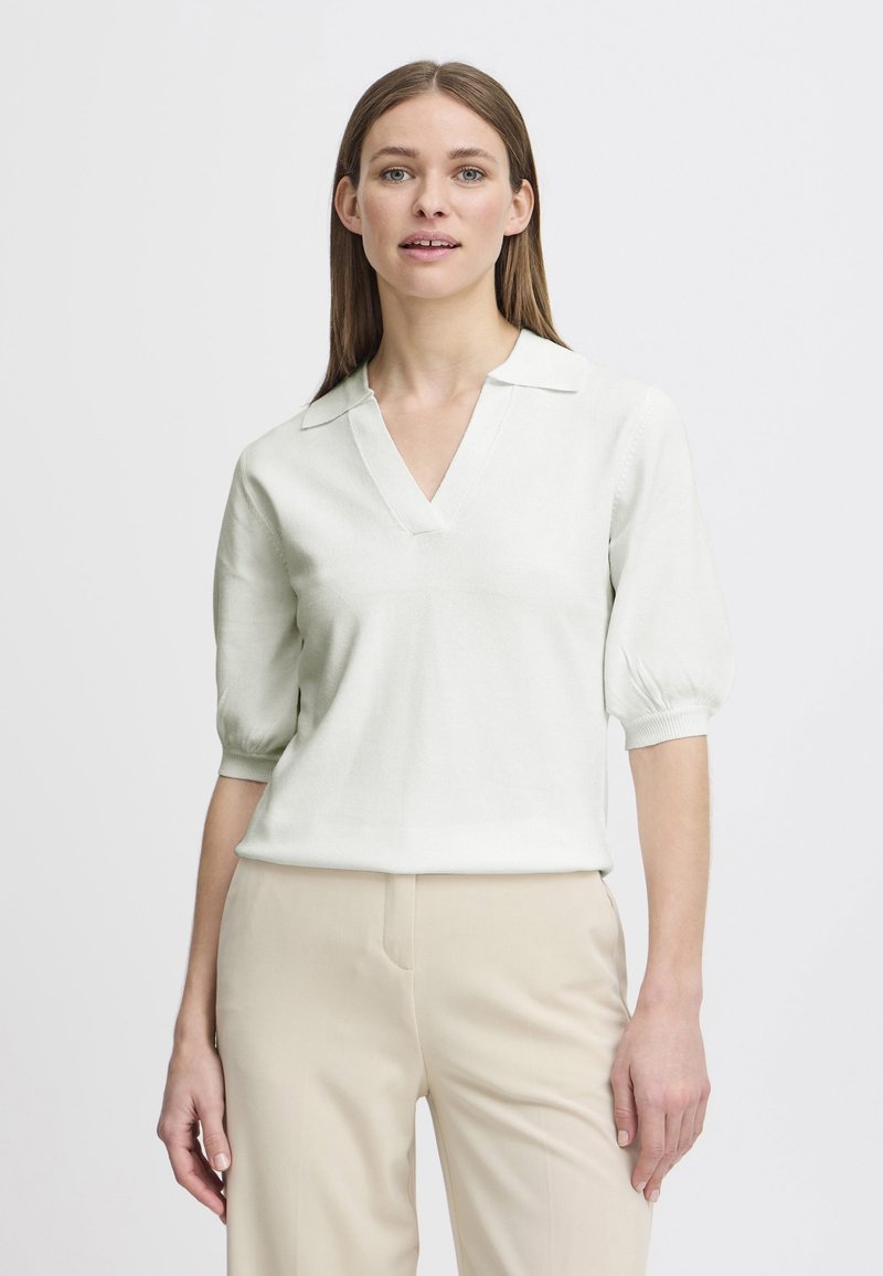 Pullover lavorato a maglia verde chiaro con scollo a V e maniche corte a sbuffo, abbinato a pantaloni beige. Texture morbida e vestibilità rilassata.