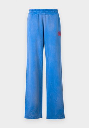 Persona che indossa una maglietta bianca a maniche lunghe con scollo a V, pantaloni larghi blu brillante con logo rosso, sneakers bianche e un berretto da baseball blu scuro, in piedi.