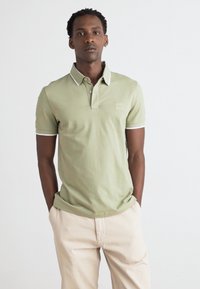 Polo vert clair avec col et finition des manches blancs, arborant un petit logo sur la poitrine. Associé à un pantalon beige, porté de manière décontractée.