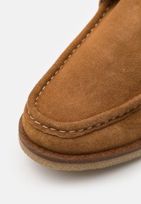 Zamszowe mokasyny w kolorze tan, z kontrastującym białym przeszyciem, zaokrąglonym czubkiem i grubą gumową podeszwą dla większej trwałości. Miękka tekstura zwiększa komfort.