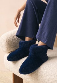 Pantofole in morbido peluche blu navy con una texture soffice e pelosa, caratterizzate da un risvolto e una punta arrotondata, posizionate su una superficie chiara e testurizzata.