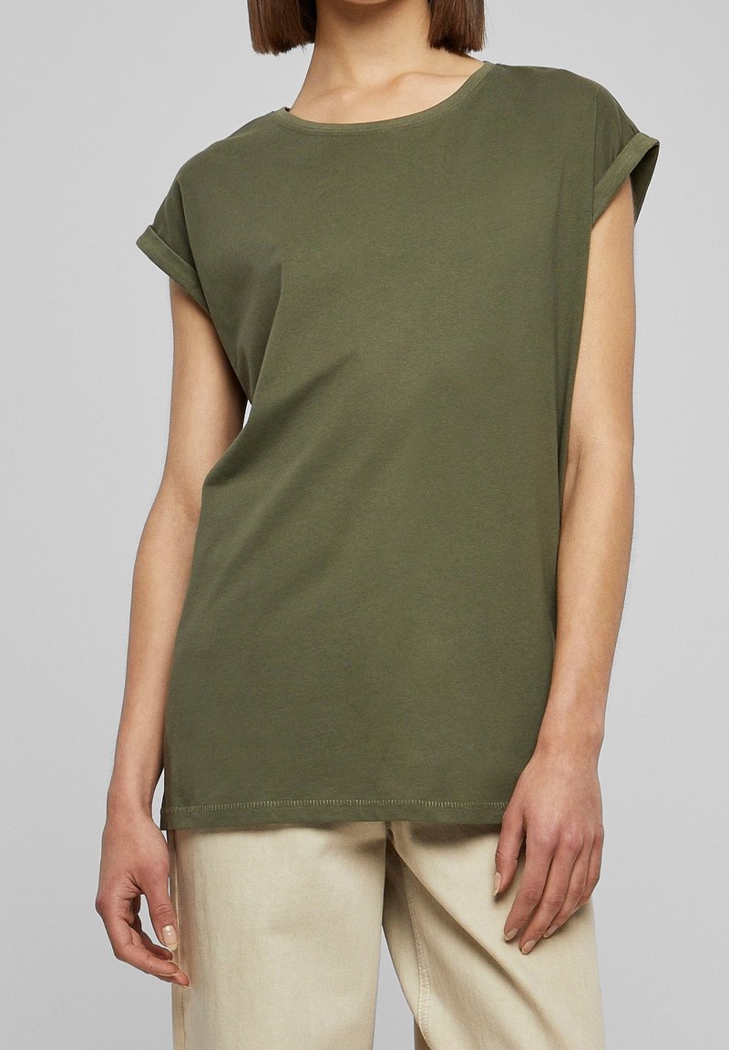 Femme portant un t-shirt vert olive à manches courtes avec revers et un pantalon beige clair sur un fond uni.