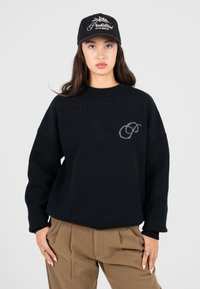 Junge Frau trägt einen schwarzen Pullover mit kleinem Logo, braune Hosen und eine schwarze Mütze mit weißem Text, vor einem einfarbigen grauen Hintergrund.
