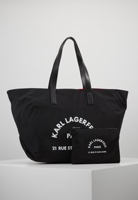 Sac fourre-tout noir et pochette assortie avec texte blanc "KARL LAGERFELD PARIS 21 RUE ST-GUILLAUME", présentés sur une surface blanche contre un fond gris.