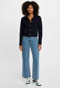 Marineblauwe, korte cardigan met gouden knopen en twee borstzakken, gedragen met lichtblauwe wijde jeans en witte sneakers met blauwe accenten.