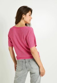 Top de punto rosa de manga corta con corte relajado, tela texturizada y acentos acanalados. Combinado con jeans de mezclilla gris claro.