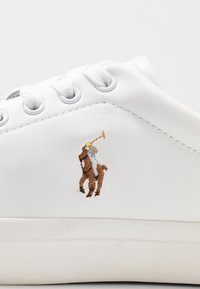 Biała skórzana tenisówka z haftowanym brązowym wzorem gracza w polo. Posiada okrągłe białe sznurówki i metalowe przelotki do zapięcia.