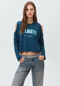 Turkizna skrajšana pulover s hladnimi rameni in napisom "SOULMATE NO MATTER WHAT". Oblikovan s hlačami z visokim pasom v modri barvi in z naglavnim pasom.