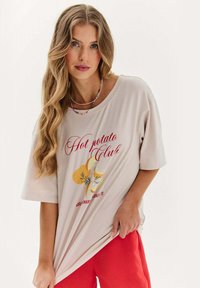 Mladá žena s dlouhými vlnitými blond vlasy má na sobě béžové oversize tričko s nápisem "Hot potato Club" a červené kalhoty na bílém pozadí.