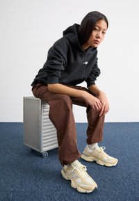 Felpa nera con cappuccio, pantaloni marroni larghi e sneakers beige con dettagli bianchi. Un contenitore di alluminio funge da seduta.