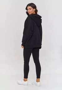 Czarna, oversize bluza z kapturem, w zestawieniu z czarnymi legginsami i białymi trampkami. Prosty design, gładka faktura, brak widocznych wzorów.