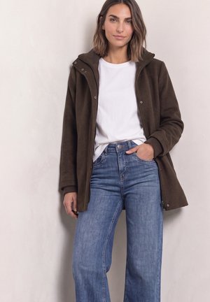 Femme debout contre un mur, portant une veste marron, un t-shirt blanc et un jean bleu, une main dans la poche, regardant la caméra.