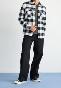 Camicia di flanella a quadri bianco e nero con due tasche sul petto, indossata aperta su una t-shirt olivastro, abbinata a jeans neri a gamba larga e sneakers nere.