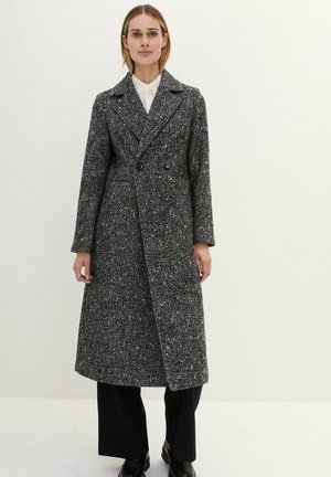 Cappotto doppiopetto in un tessuto testurizzato nero e grigio, con una silhouette lunga, colletto a punta e tasche laterali. Pantaloni e scarpe neri.