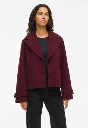 Veste en mélange de laine bordeaux avec un large col, deux poches avant et des boutons noirs. Présente des manches raccourcies avec détail aux poignets.