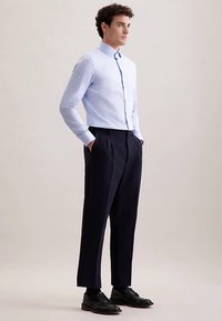 Camicia celeste chiusa con bottoni e collo con risvolto, abbinata a pantaloni eleganti blu navy a gamba stretta. Il modello è in piedi con le mani in tasca, indossando scarpe nere.