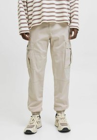 Beige Cargohose mit elastischen Bündchen, mehreren seitlichen Taschen und glatter Textur, kombiniert mit hellen Sneakers.
