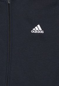 Vêtement noir zippé avec un logo blanc Adidas sur la poitrine, avec trois bandes diagonales au-dessus du nom de la marque.