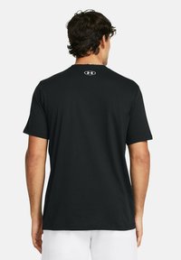 Vīrietis valkā vienkāršu melnu T-kreklu ar īsām piedurknēm, uz kura aizmugures pie kakla ir neliels Under Armour logo, apvienojot to ar baltām šortiem.