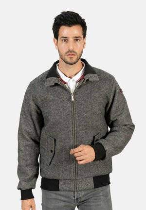 Veste en chevrons grise avec un col noir, fermeture éclair et poches avant. Dotée de poignets et d'un ourlet côtelés, portée sur un polo blanc.