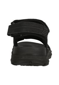 Mustat sandaalit, joissa on texturoitu hihna ja säädettävä kiinnitys. Ominaisuuksiin kuuluu paksu kumipohja mukavuutta ja kestävyyttä varten. Taustakuva.