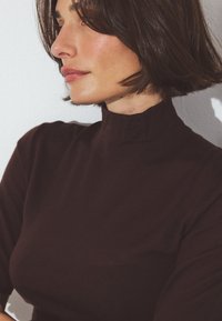 Ruskea korkeakauluksinen paita, joka on valmistettu pehmeästä materiaalista. Paidassa on lyhyet hihat ja se istuu hyvin vartalonmyötäisesti. Taustalla on vaalea seinä.