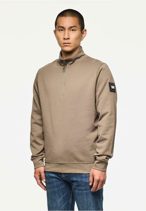 Bruine ritssluiting sweatshirt met een hoge kraag, ribgebreide manchetten en een textuurpatch op de linker bovenarm. Gedragen met een blauwe spijkerbroek.