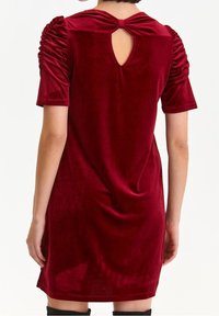 Vista posteriore di un vestito in velluto a maniche corte di un rosso intenso con spalle arricciate, un taglio a chiave e un fiocco di stoffa sul collo.