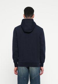 Námornícky modrá mikina s kapucňou na šnúrku, z jemného materiálu, s pružnými manžetami a klokanie vrecko, v kombinácii so svetlými džínsami.