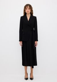 Černý dlouhý blazer s obvodovým designem, zahrnuje detail s mašlí a plisovanou černou sukni. Oblečení doplňují černé lodičky s špičatou špičkou.