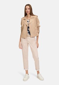 Veste en suédine beige avec un col à revers, associée à un haut à imprimé floral, un pantalon à ourlet roulé rose clair et des baskets blanches.