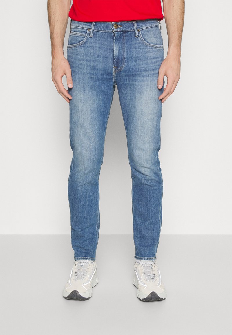 Jeans de mezclilla azul claro con un diseño ajustado, que cuentan con cinco bolsillos y un desvanecimiento sutil. Combinados con zapatillas deportivas de color claro.