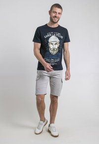 T-shirt marine avec un graphique d'un homme barbu et des rayures, associé à un short cargo gris clair. Baskets blanches avec des accents marron.