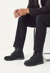 Schwarze Wildleder-Stiefeletten mit Schnürdesign, seitlichem Reißverschluss und robuster Sohle, kombiniert mit schwarzen Hosen. Die Stofftextur ist sichtbar.