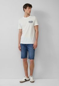 T-shirt en coton blanc avec un imprimé graphique bleu sur la poitrine, associé à un short en denim bleu et des baskets blanches avec des accents noirs.