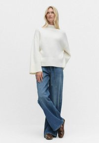 Vit oversized tröja med ribbade ärmslut och nederkant, matchad med vida blå jeans och bruna skor, stående på en neutral bakgrund.