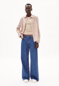 Camicia a righe rosa e bianca, canotta aderente beige e jeans in denim blu a gamba larga con cordino, abbinati a scarpe bianche.