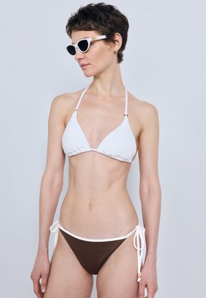 Femme aux cheveux courts et foncés, portant des lunettes de soleil blanches et un bikini bicolore avec un haut blanc et un bas marron à nouer sur les côtés, regardant vers la gauche.