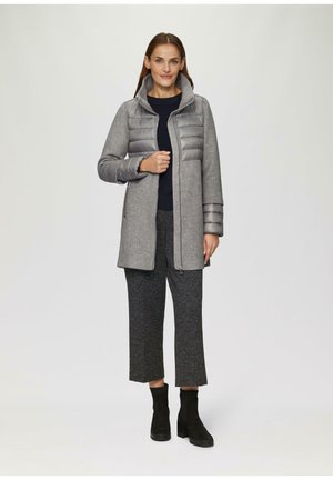 Manteau en mélange de laine gris avec une partie supérieure matelassée et une capuche grises. Porté par-dessus un pull noir et un pantalon court texturé gris, accompagné de bottes noires.