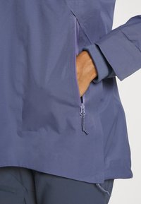 Granatowa wodoodporna kurtka ma boczną kieszeń na zamek z purpurowymi detalami oraz rękaw zakończony mankietem o teksturowanym wykończeniu.